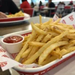 A close-up shot of a serving of Steak 'n Shake’s freshly fried shoestring fries, golden and crispy, piled high in a red-and-white classic fast-food tray. The fries glisten slightly, showing their rich, tallow-fried texture, with a small container of ketchup on the side. The background is blurred, with a casual dining setting. The lighting is natural, and the image has an amateur feel, as if taken with an iPhone 15 Pro and shared on Reddit. No text, no watermarks, just an appetizing, real-world shot