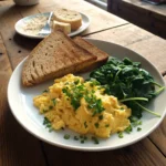 A plate of golden scrambled eggs garnished with fresh parsley, served with whole-grain toast and sautéed spinach on a rustic wooden table, photographed in natural morning light.